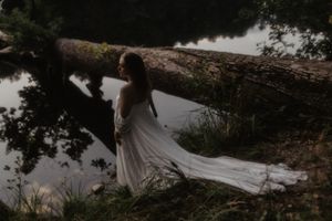 Figure in flowing white dress balanced on fallen tree over water.