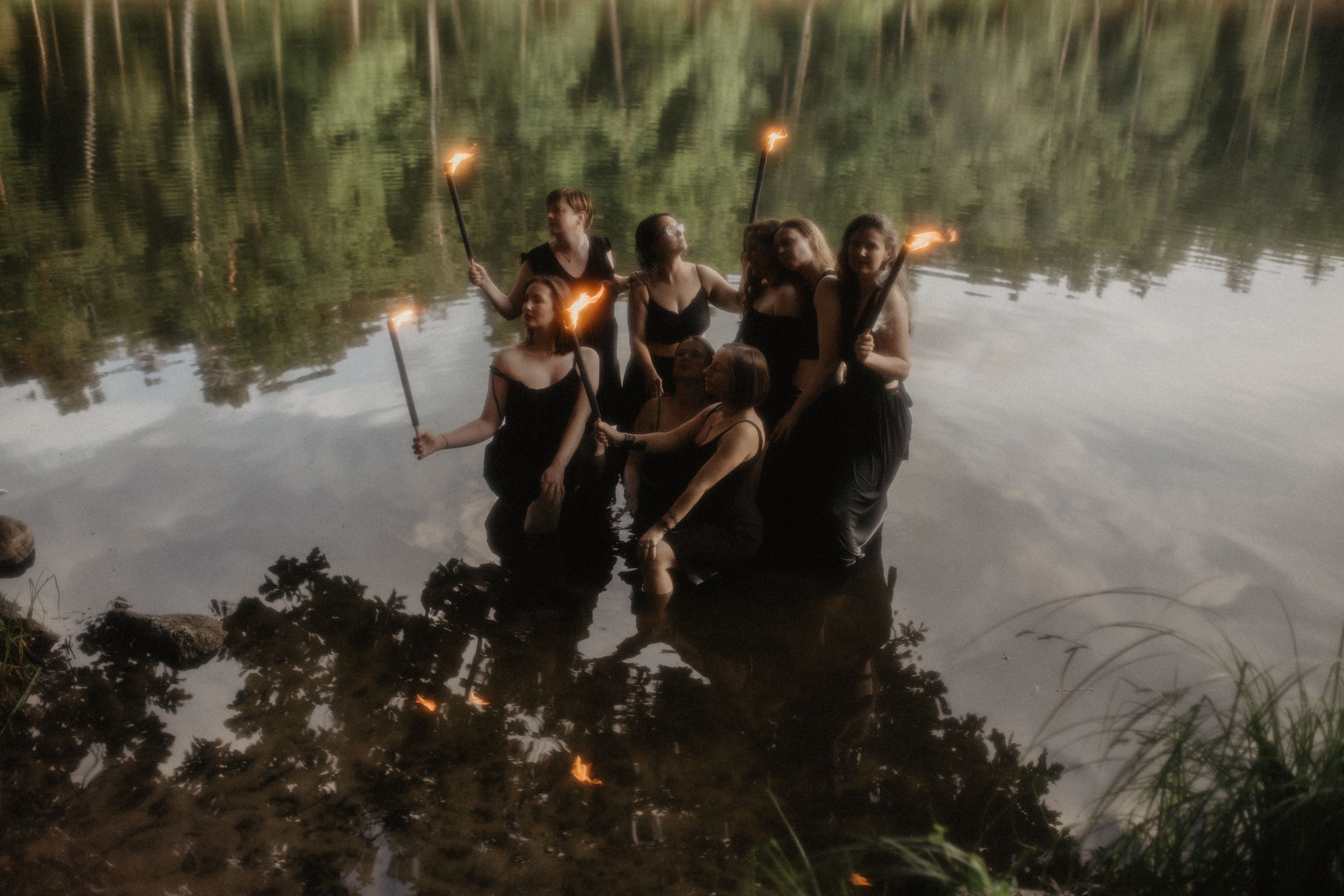 Four women holding torches stand in a misty lake surrounded by lush green trees at dusk.