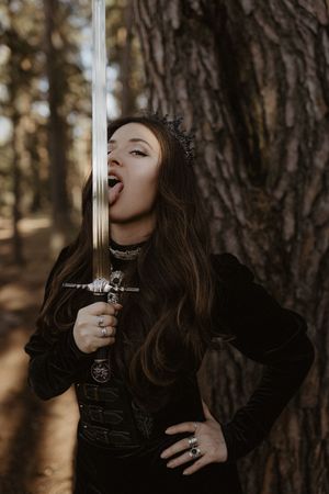 Woman in black armor costume licking a decorative sword blade in a forest setting.