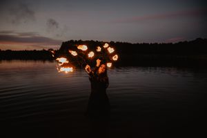 Person performing fire dancing in a lake at dusk, holding multiple flames against a twilight sky.