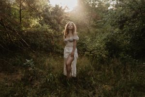 Figure in white dress standing in golden sunlit forest clearing at dusk.