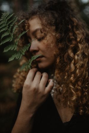 Close-up portrait with curly hair and fern leaves in moody forest setting.