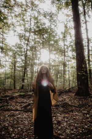 Figure in dark dress standing among tall trees with ethereal sunburst effect.