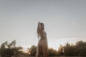 A ethereal figure in a flowing white dress stands against a misty sunset backdrop in nature.