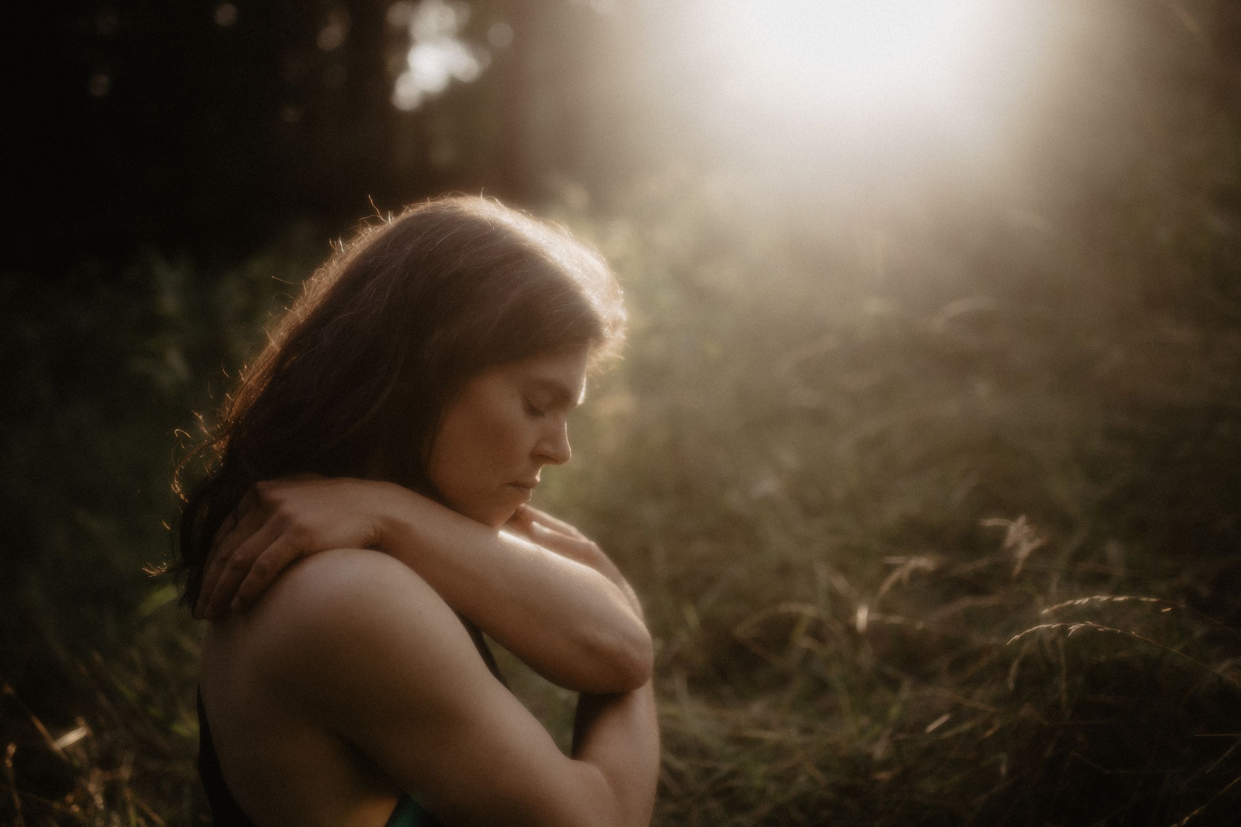A woman with brown hair hugs herself in a sunlit forest, eyes closed in a peaceful, introspective moment.