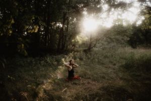 Person practicing yoga in a sunlit forest clearing surrounded by lush green trees and tall grass.