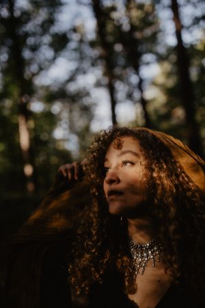 Moody portrait with golden sunlight filtering through trees highlighting curly hair and jewelry.