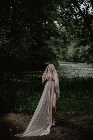 Mysterious veiled figure in white stands along forest path near water.