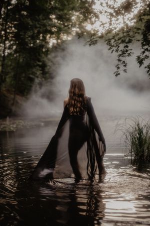 Dramatic silhouette in flowing black dress wades through misty lake at dawn.