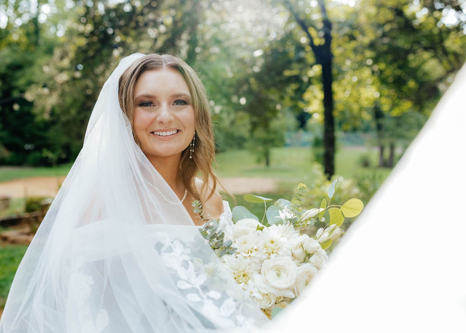 Bride holding white bouquet in outdoor garden setting with natural sunlight.