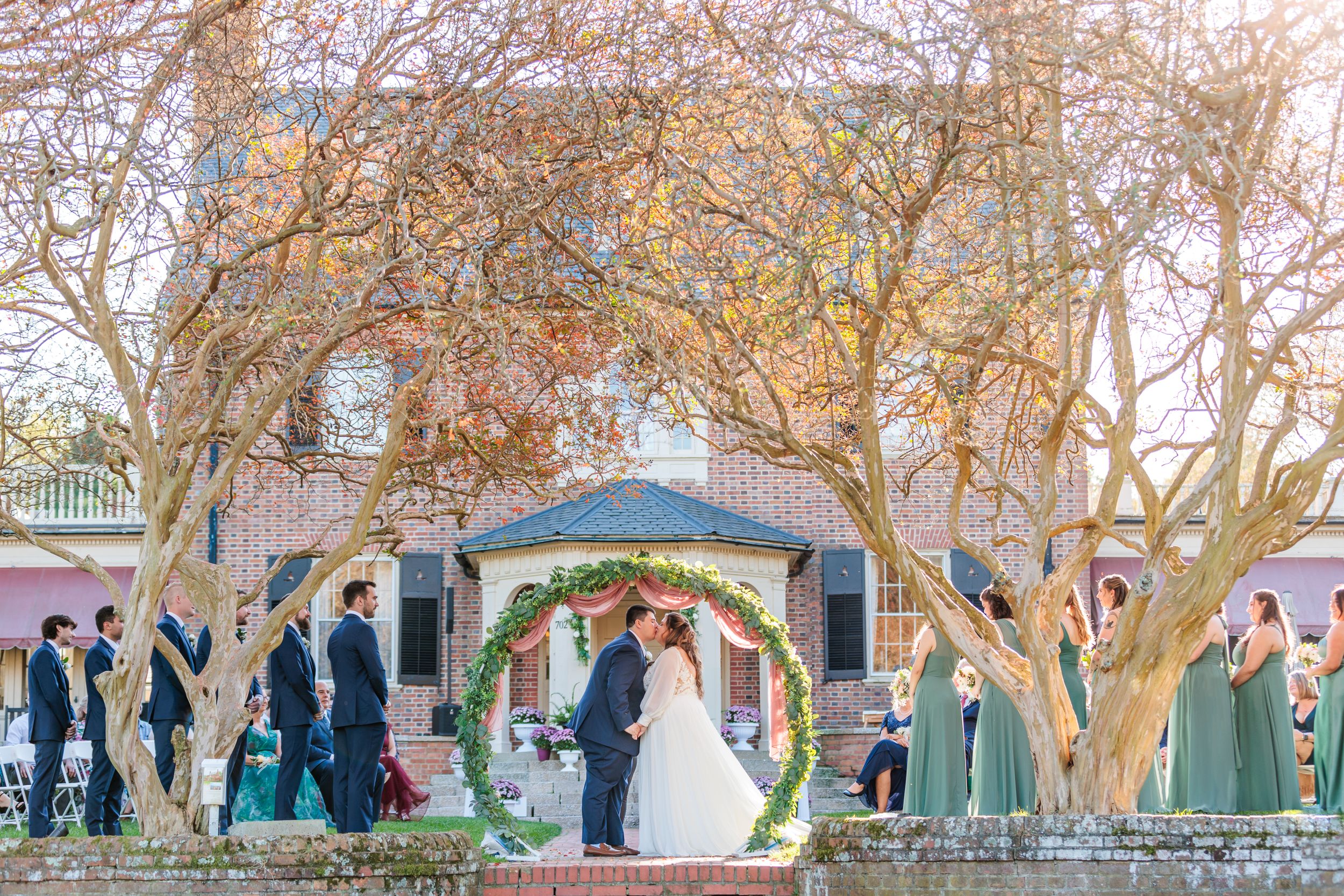 Wedding ceremony under an archway framed by twisted trees with bridal party in blue and green.