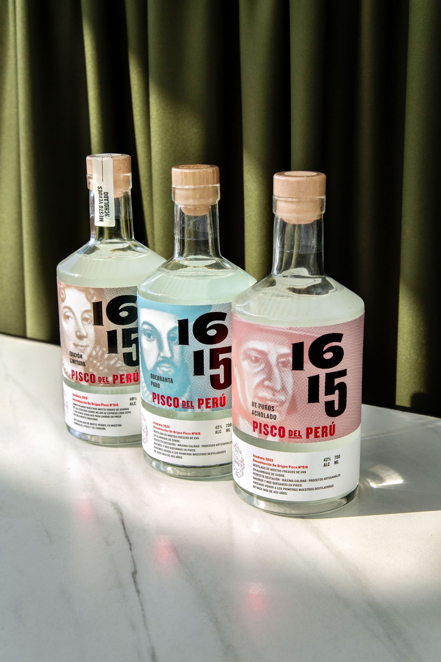 Three bottles of 1615 pisco liquor from Peru arranged on a white surface against a green curtain backdrop.