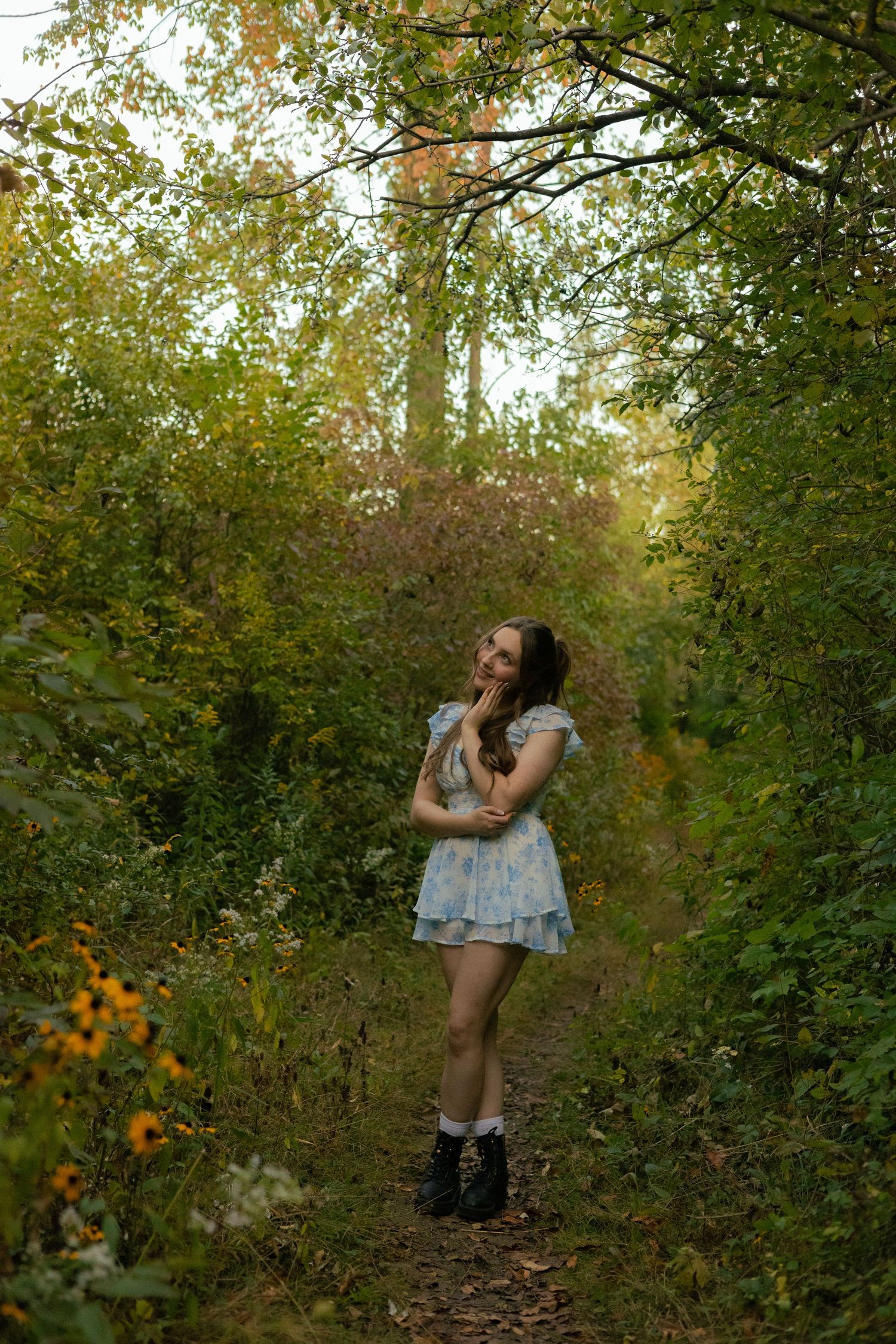 A woman in a coral dress poses gracefully among autumn leaves and trees in a sunlit forest setting.