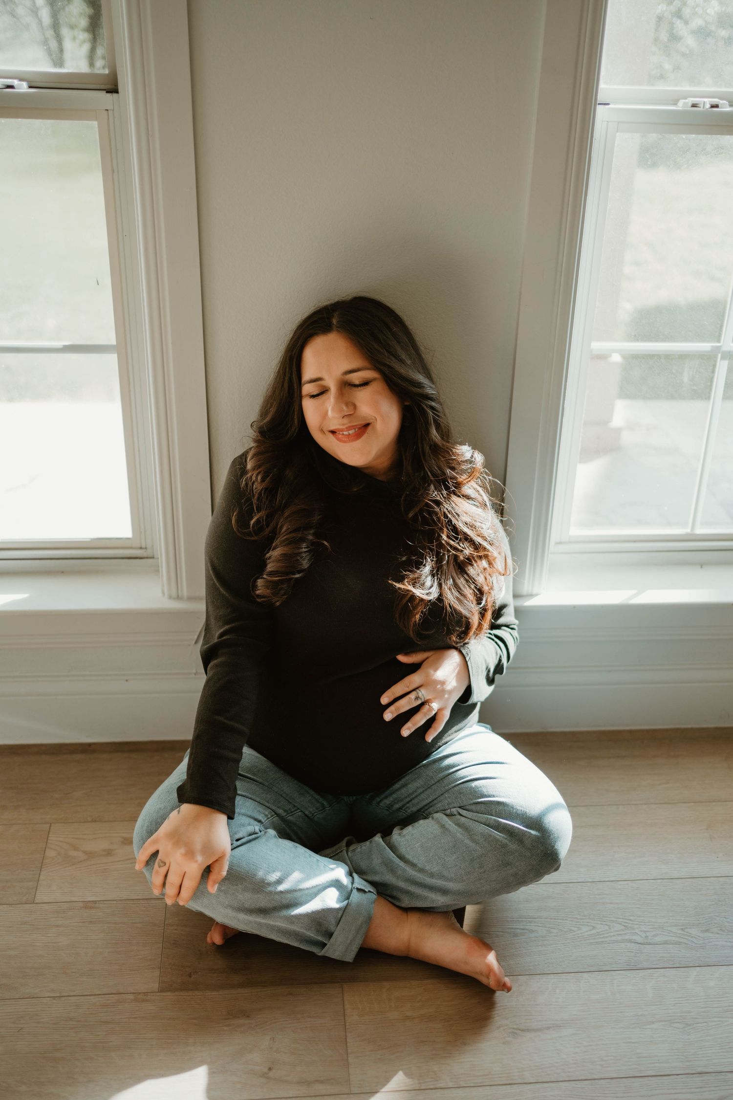 Happy person in a black sweater sits cross-legged on hardwood floor by sunny windows.