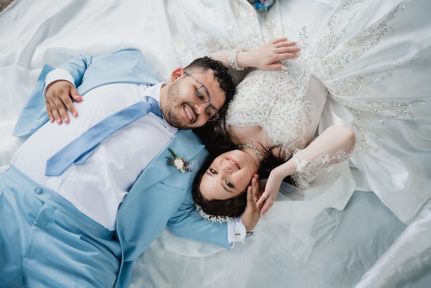 Overhead wedding photo with couple lying on white dress fabric.