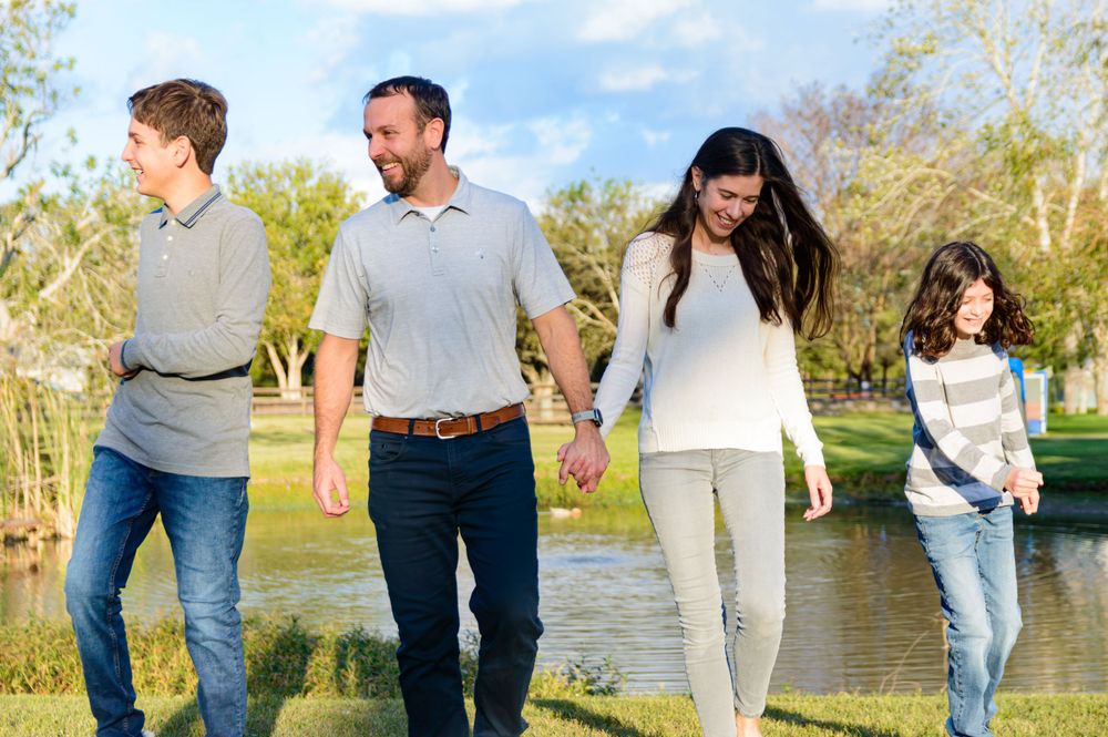 A family of four walks together holding hands along a pond's edge on a sunny day.