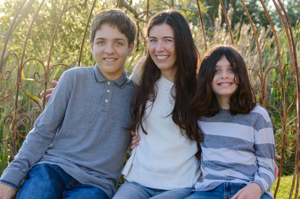 Three siblings sit together outdoors smiling against reeds and natural scenery.