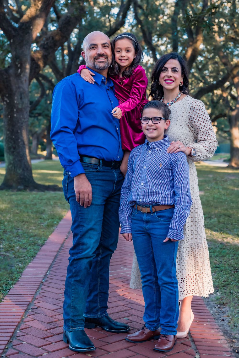 A family of four poses together on a brick path in a park setting with trees in the background.
