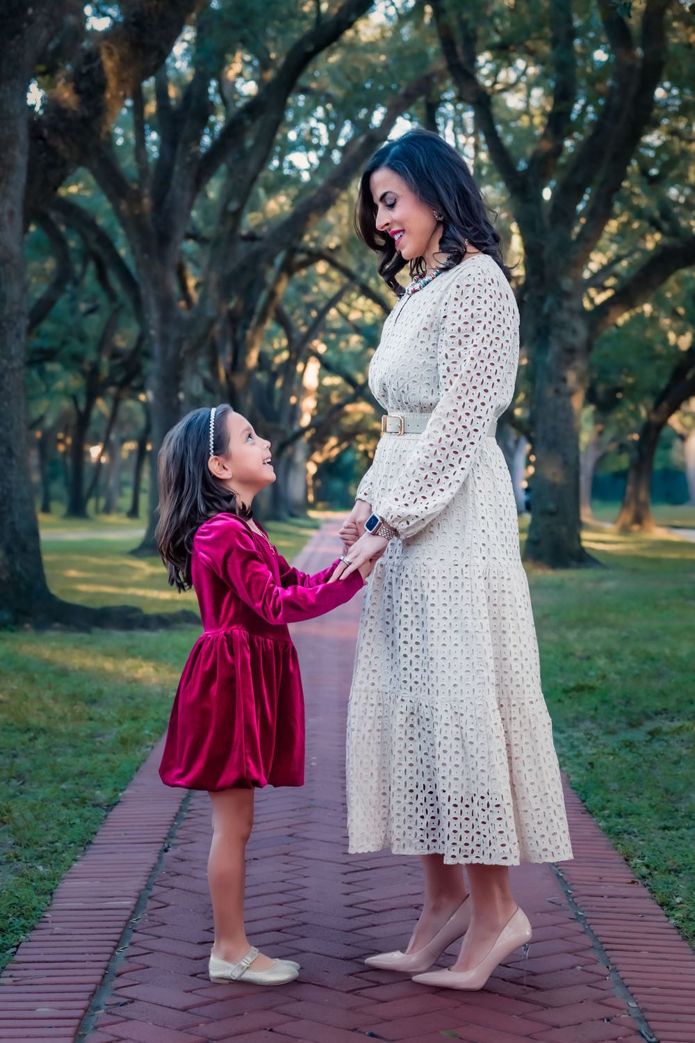 A child in a red velvet dress interacts playfully with someone wearing a white eyelet dress on a brick path.