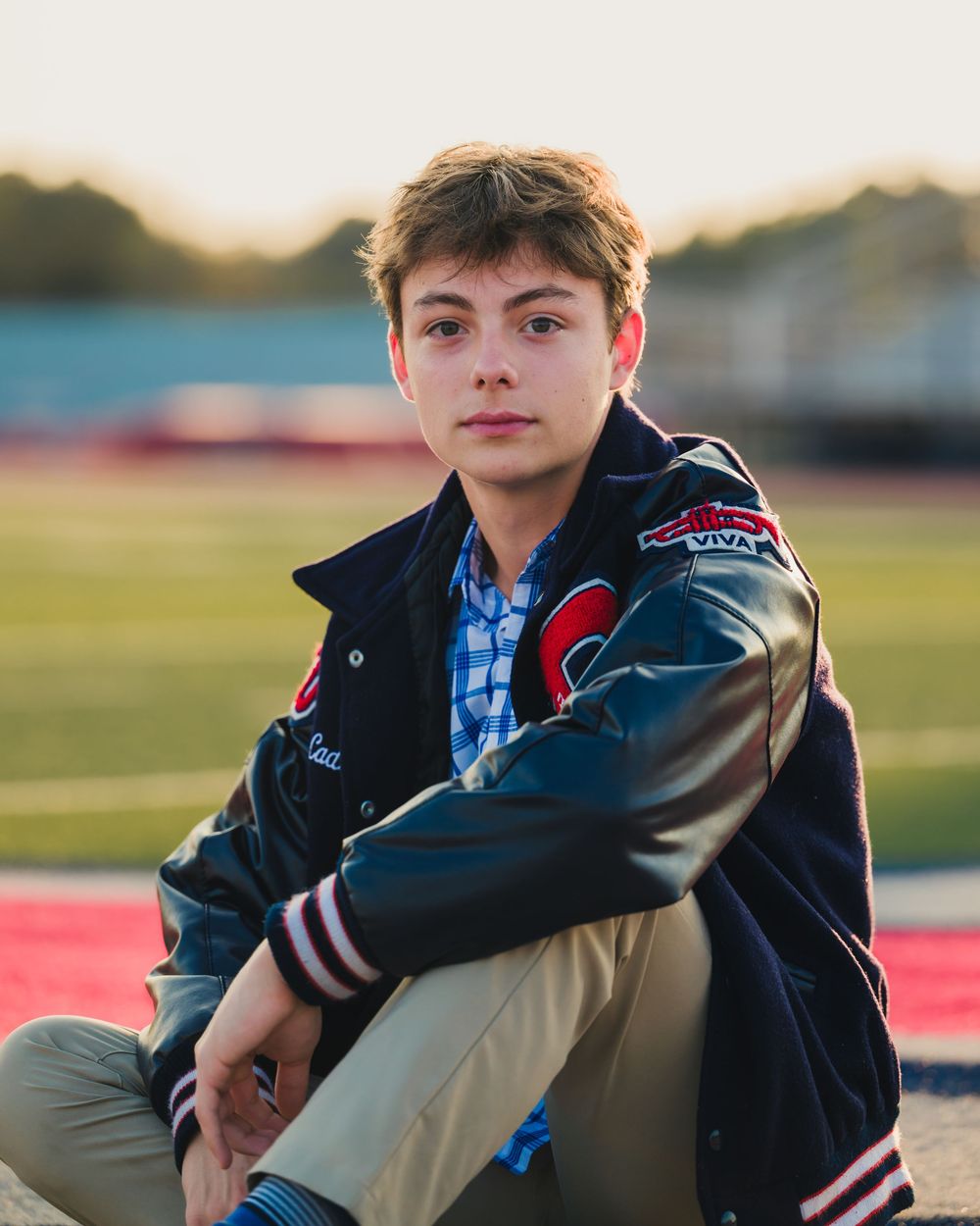 Student wearing varsity jacket and plaid shirt poses outdoors at sunset on school grounds.