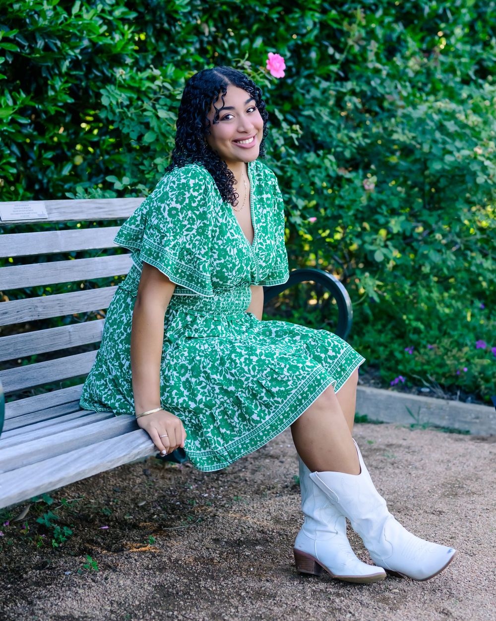 Person in a green floral dress poses on a wooden bench in a garden setting while wearing white cowboy boots.