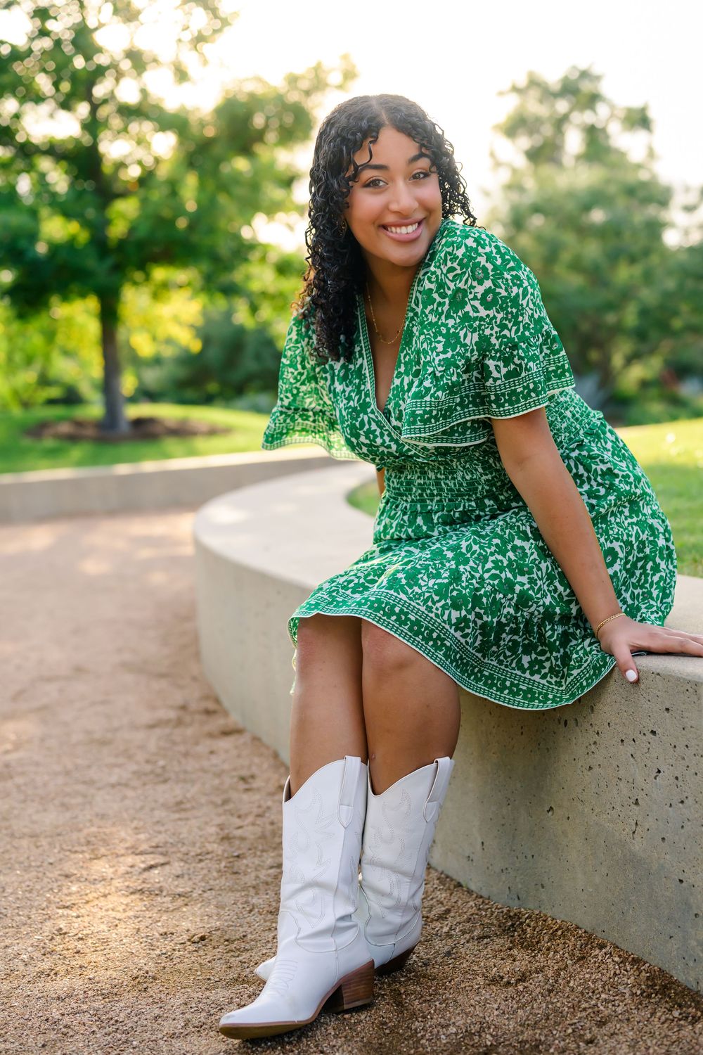 A person wearing a green floral dress and white cowboy boots sits on a concrete ledge in a sunlit outdoor setting.