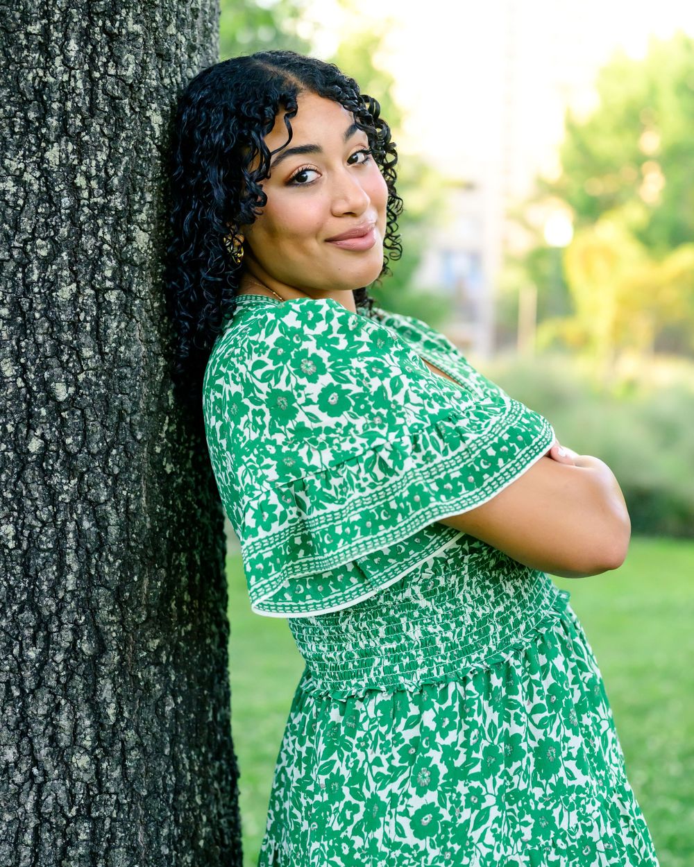 A person wearing a floral green dress poses against a tree trunk in an outdoor setting with soft natural lighting.