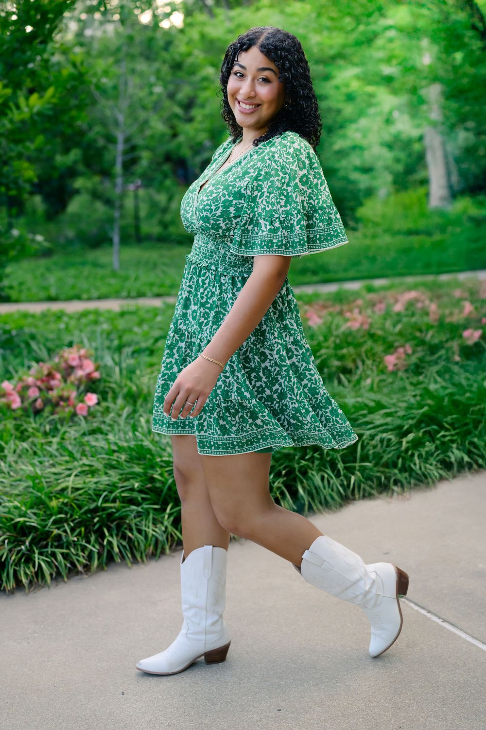 Person in a green floral dress and white cowboy boots poses on a garden path surrounded by greenery.