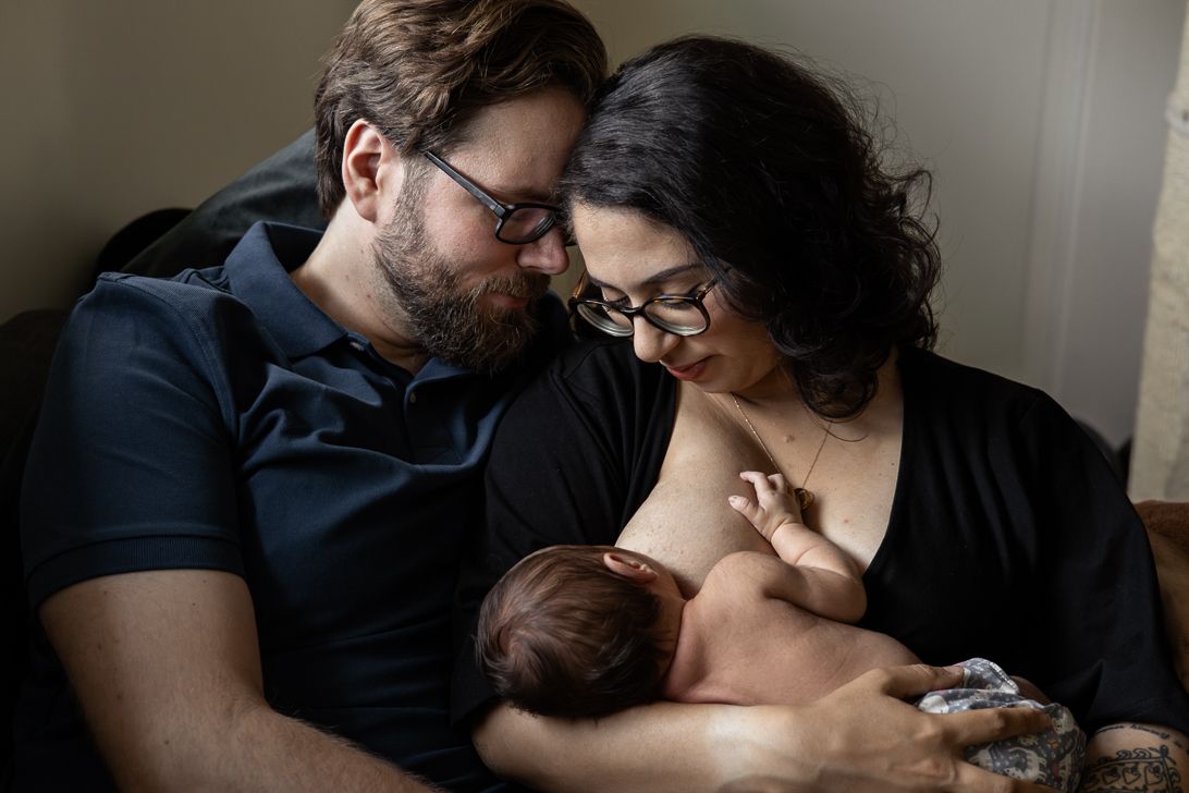 A tender family moment as parents lovingly watch their newborn baby nursing in soft natural light by a window.