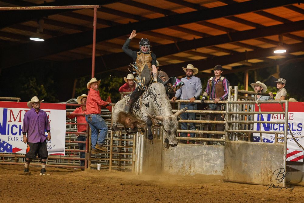 Cowboy riding bucking bronco at indoor rodeo arena at night, with spectators and handlers watching nearby.