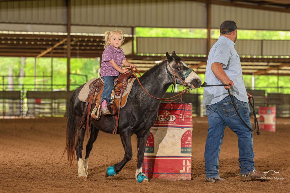 Young girl riding a black horse led by a man in an indoor rodeo arena with a red barrel nearby.