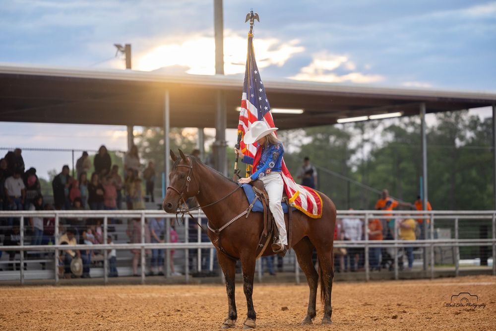 A performer rides a brown horse while holding an American flag at sunset. Jasper County Cowboy Church. Jasper Pro Rodeo