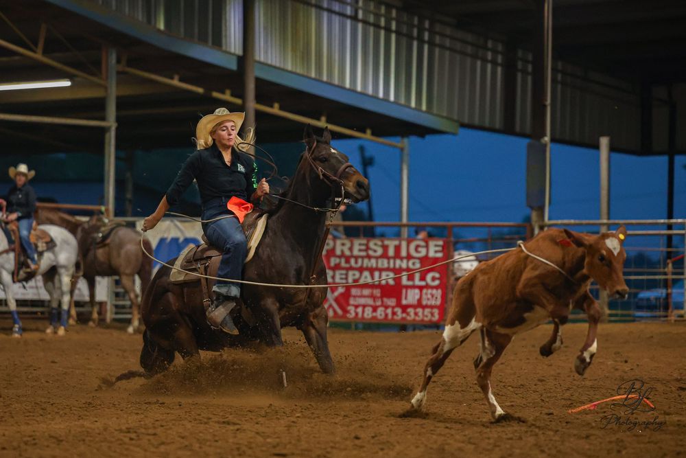 Cowgirl on horseback competing in a rodeo event at an indoor arena, chasing a calf on dirt ground.