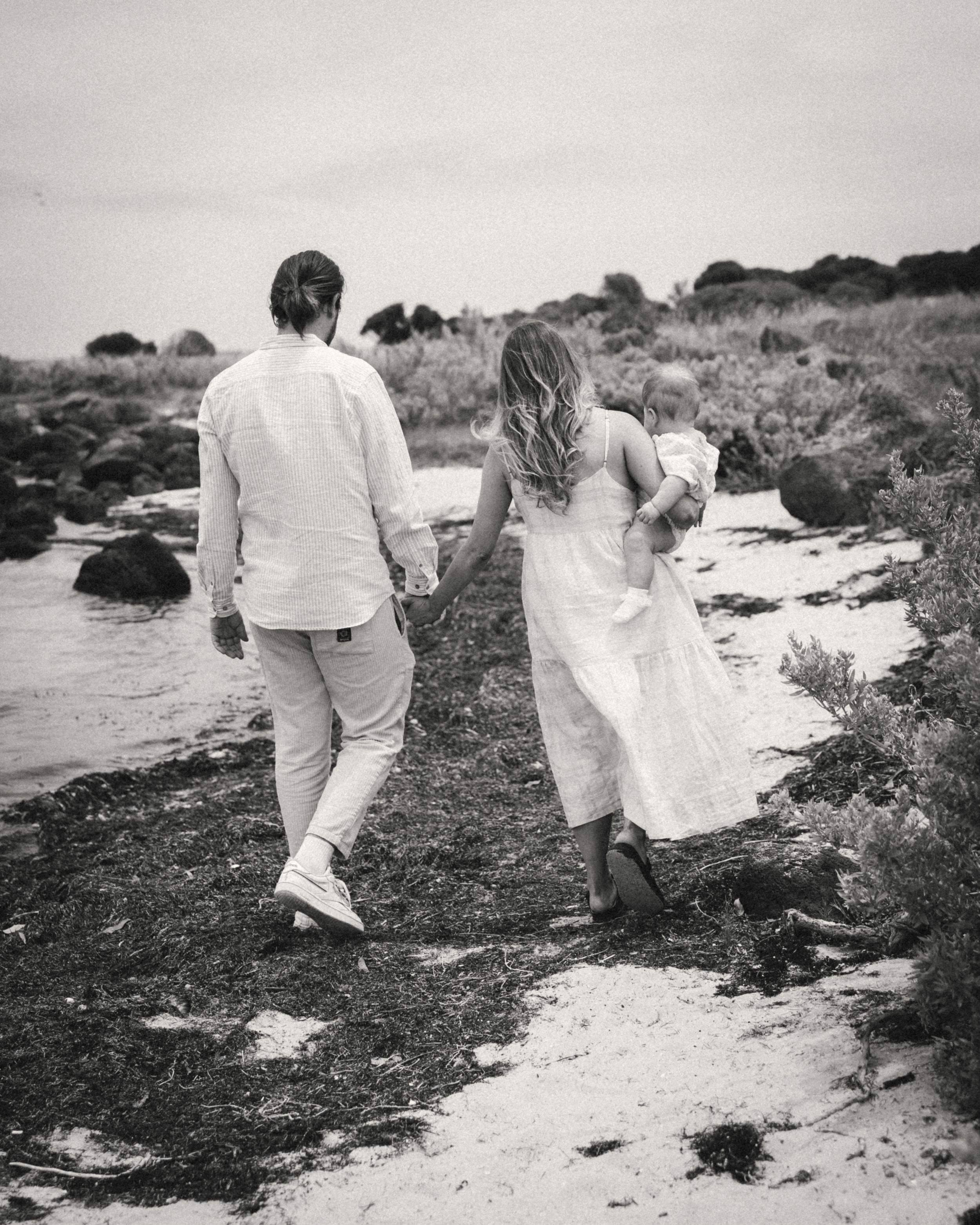 A couple and their baby walks along a rocky coastal path in black and white photography.