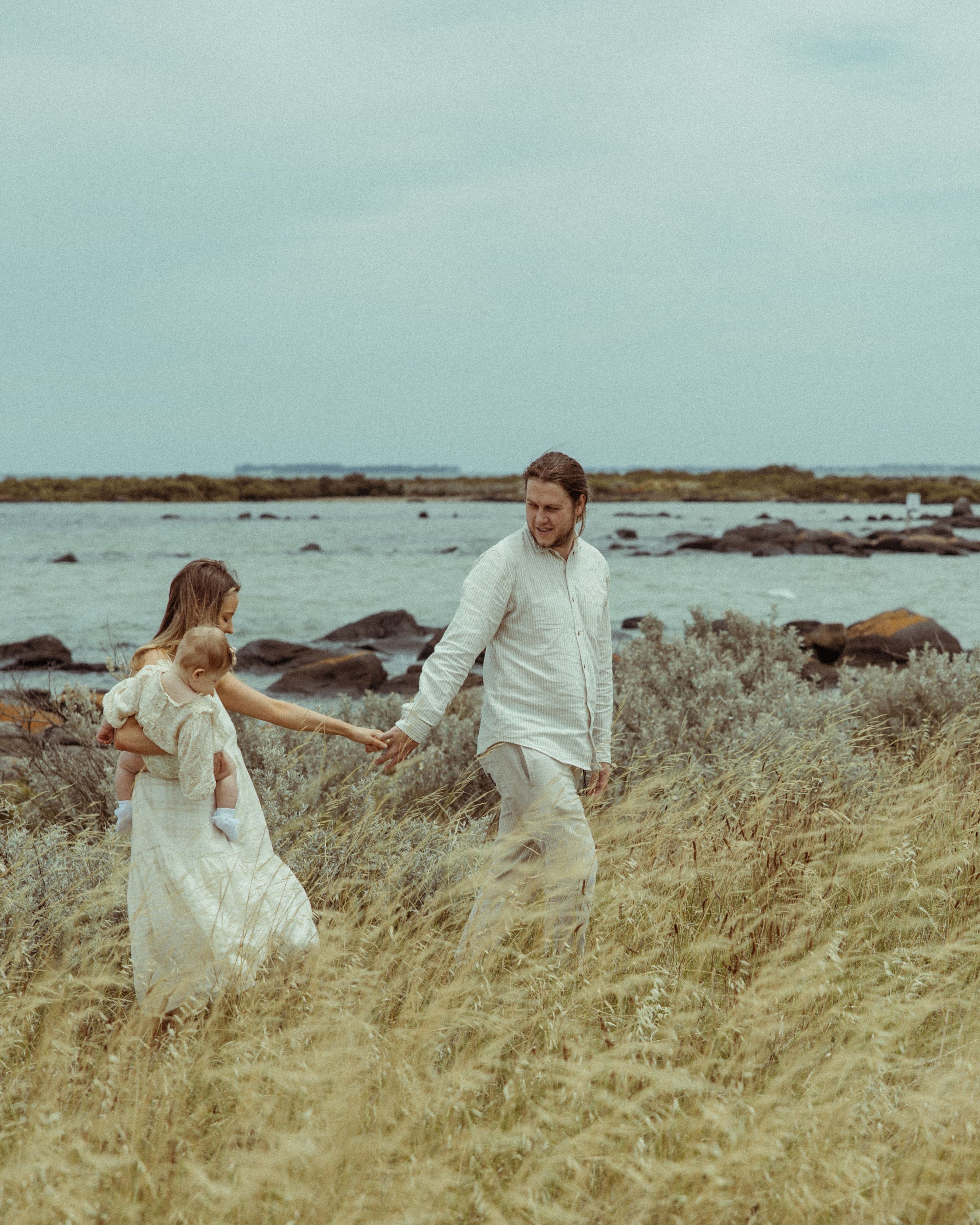 Couple walking together with their daughter through tall grass near a rocky beach on a cloudy day.