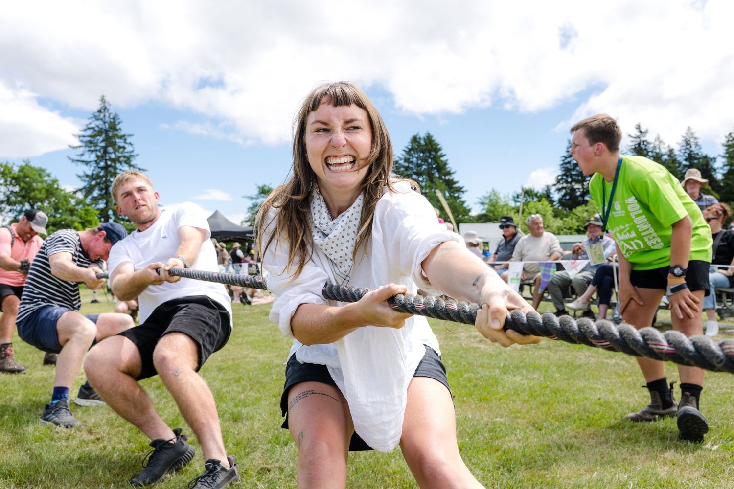 Group of people playing tug of war outdoors on a sunny day with trees in the background.