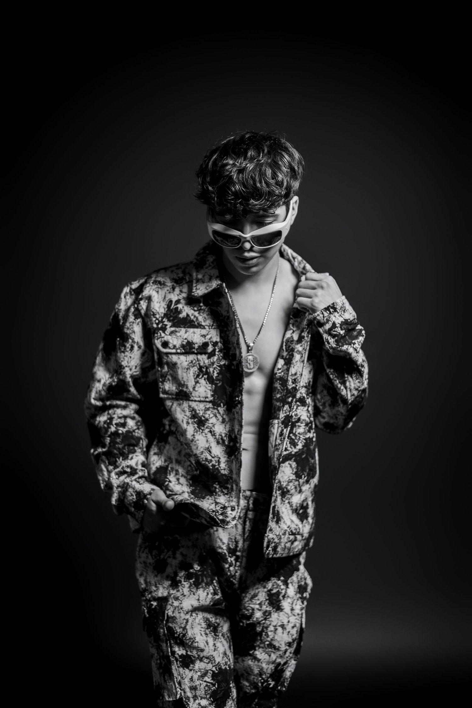 Artistic portrait of person in camouflage jacket and sunglasses against dark backdrop.