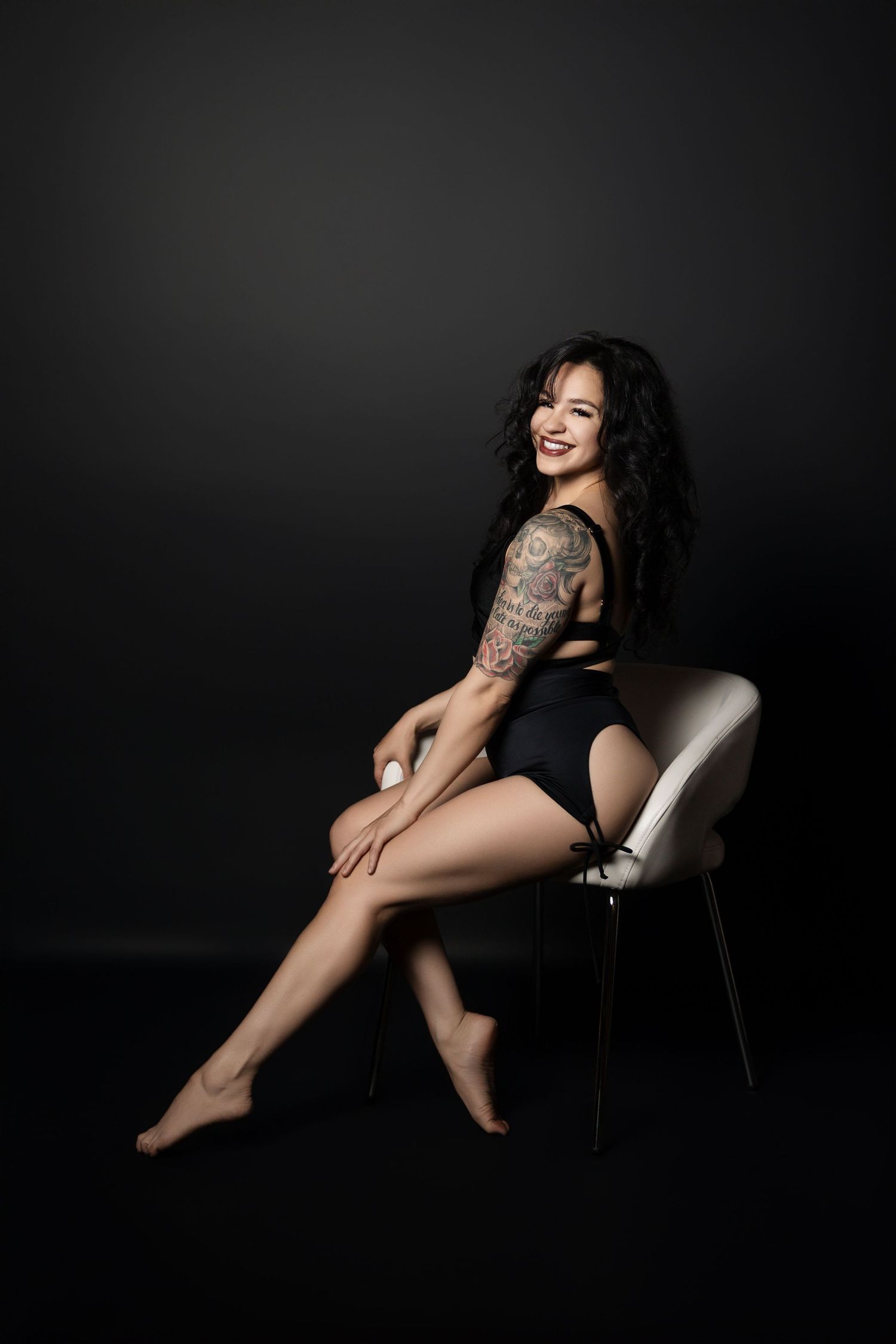 Model with tattoos poses elegantly on white chair against dark background.