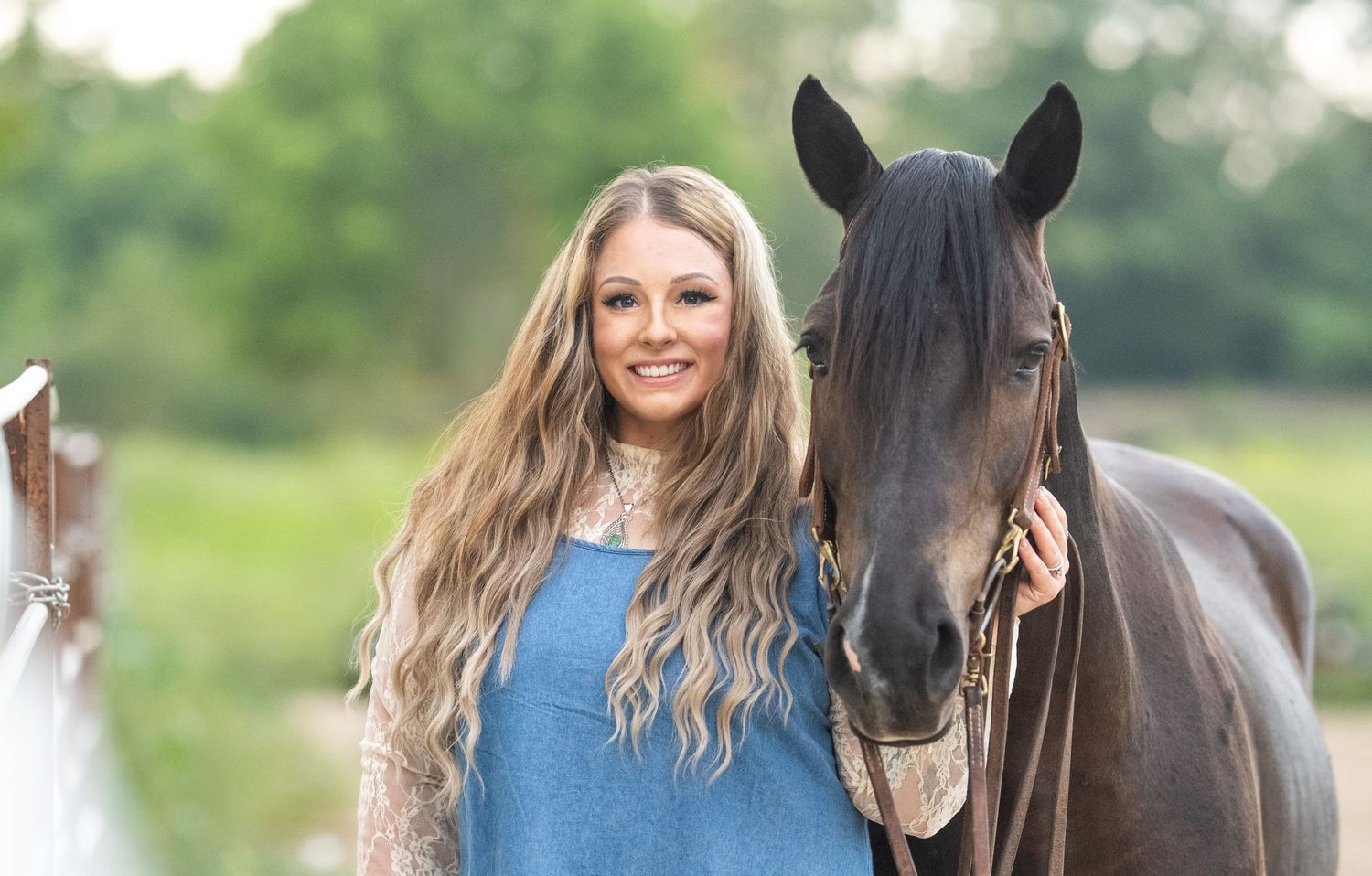 Dark brown horse with bridle poses with someone in denim and white lace top in grassy field.