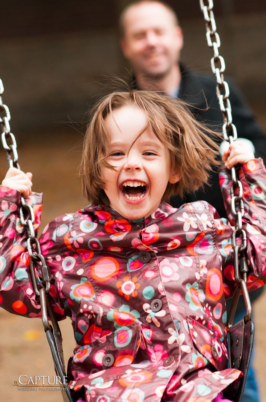 A child in a floral patterned jacket laughs joyfully while swinging outdoors.