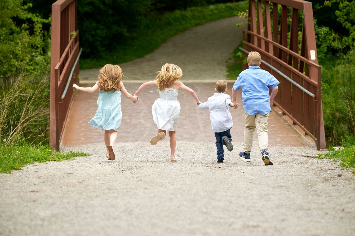 Children run across a wooden bridge on a gravel path through a green forest area.