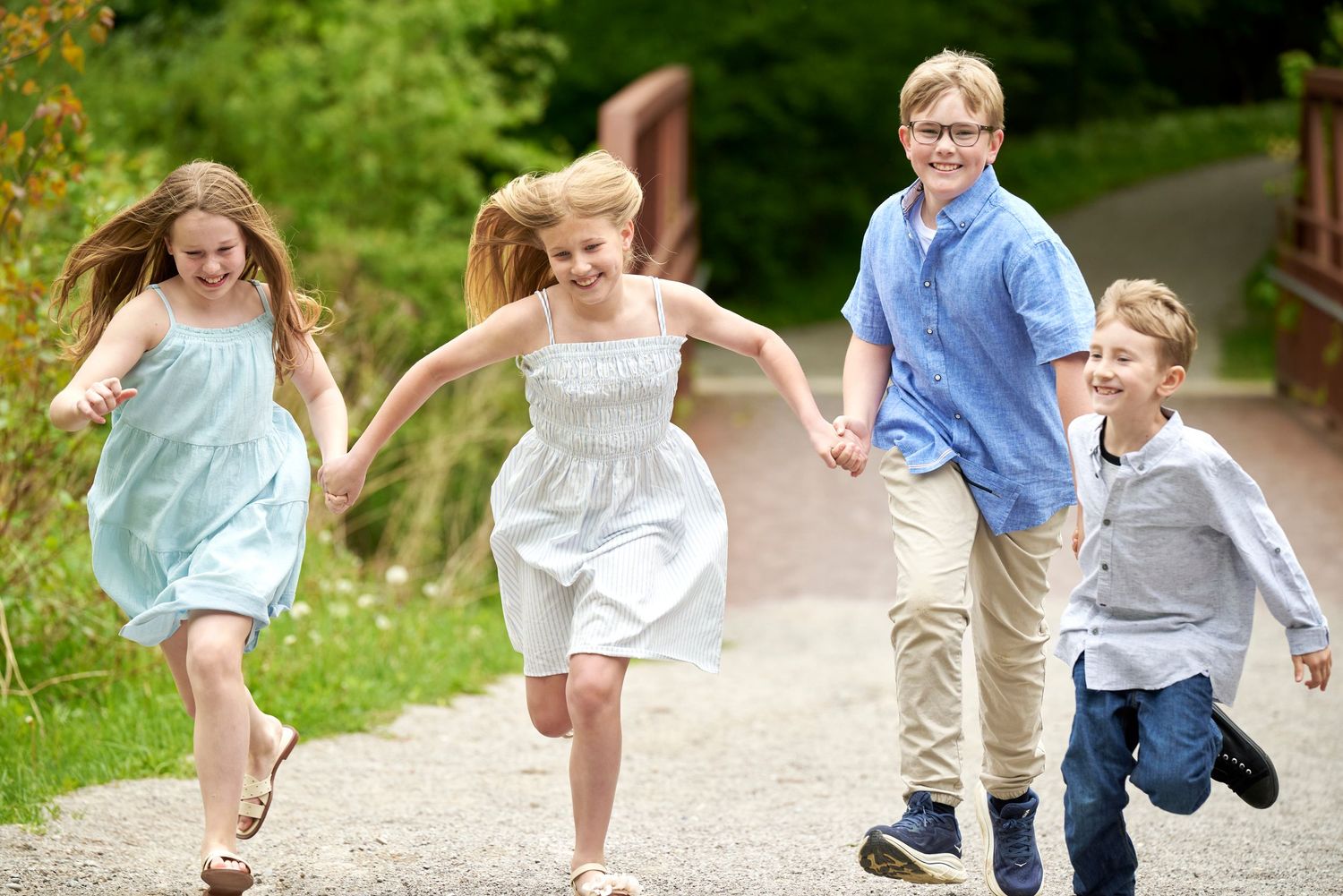A sequence of joyful people running and playing together along a tree-lined path in a summer garden setting.