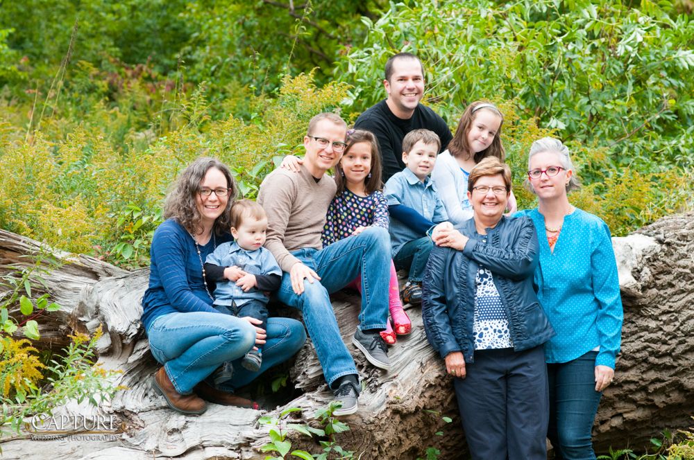 Large family group sitting together on fallen tree trunk in wooded outdoor setting.