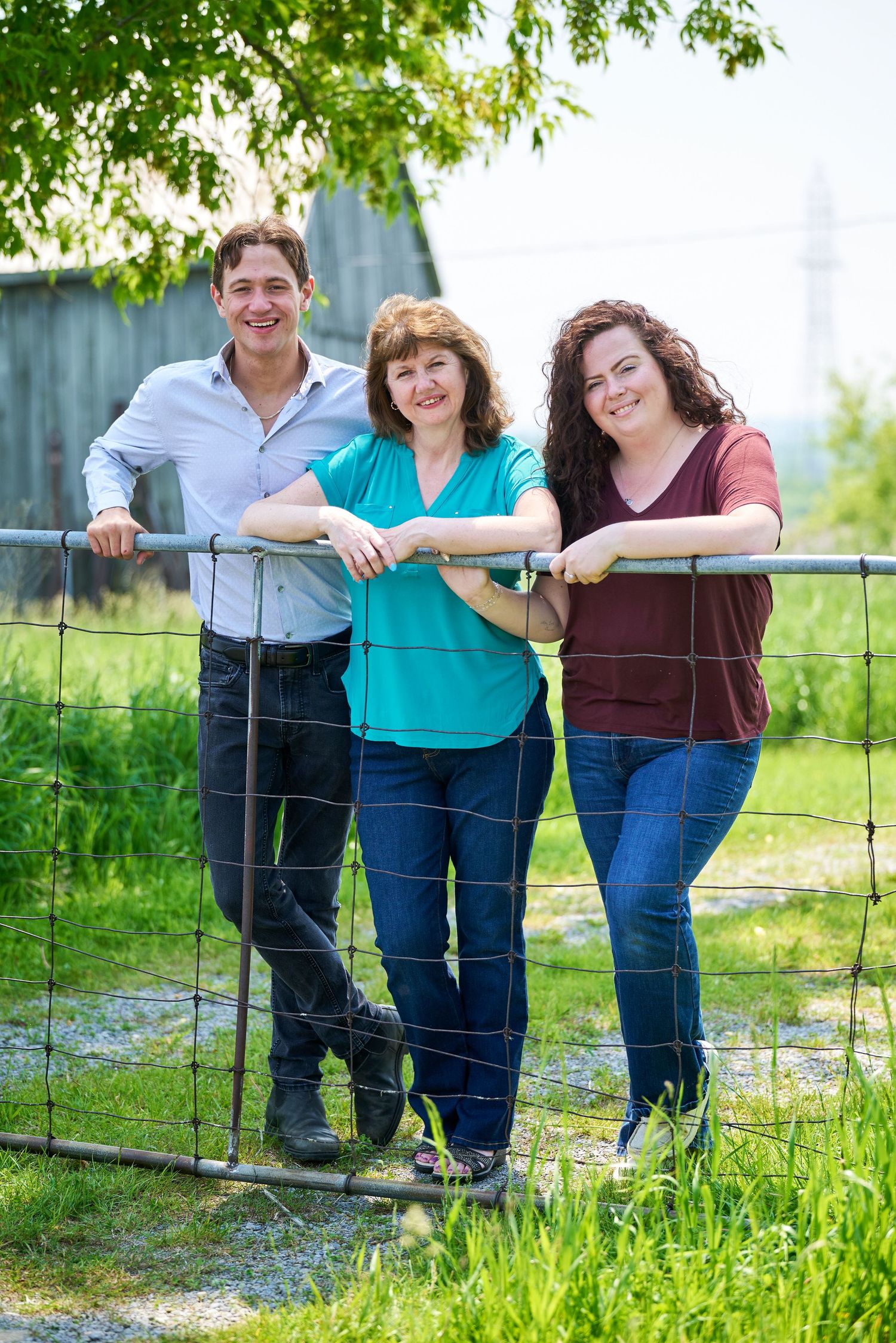 Family poses together by rustic fence in outdoor setting with green grass and trees in the background.
