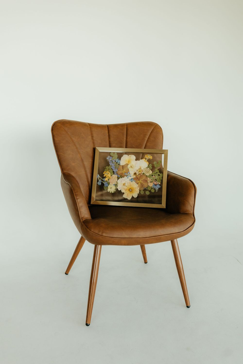 Yellow and white pressed flower art in gold frame resting on brown velvet chair, white background.