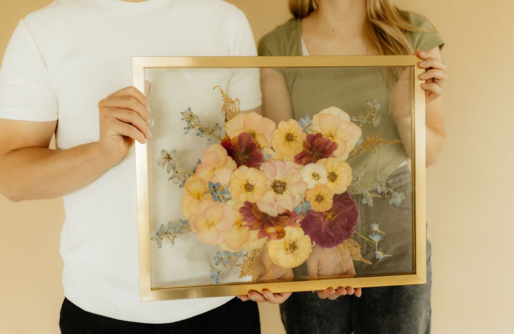 Couple holding a large gold-framed pressed flower bouquet preservation artwork together.