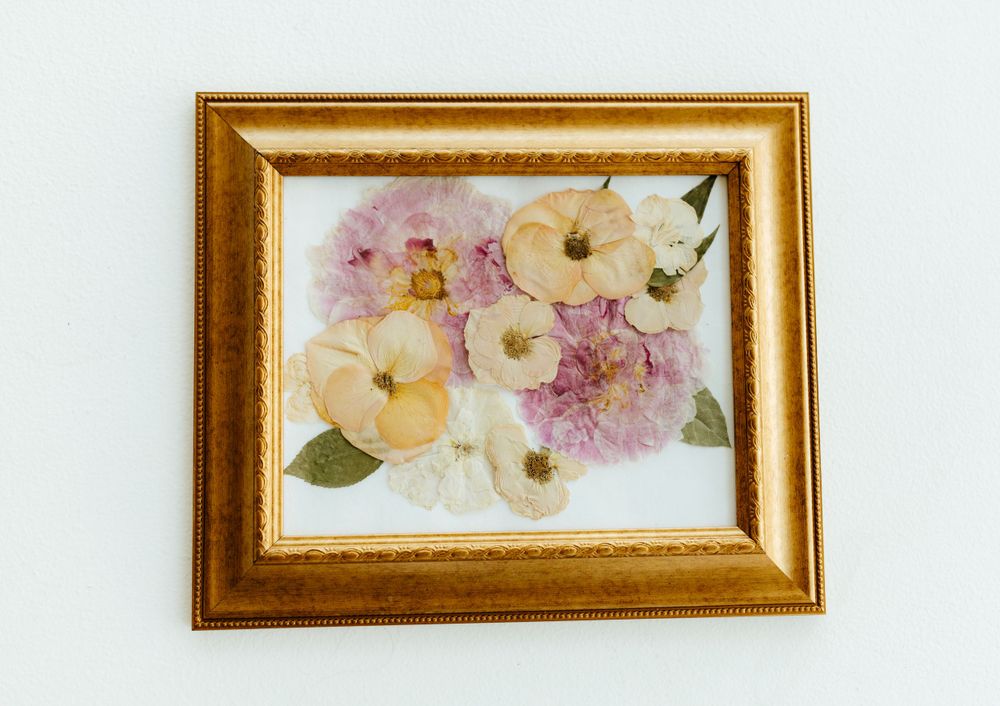 Framed pressed flower art featuring pink, peach, and white blooms in an ornate gold frame.