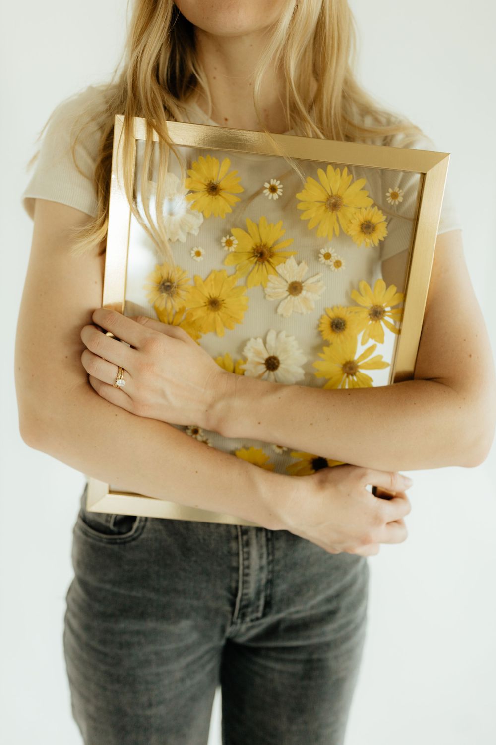 Woman holding a gold frame with pressed yellow and white daisy flowers on clear background.