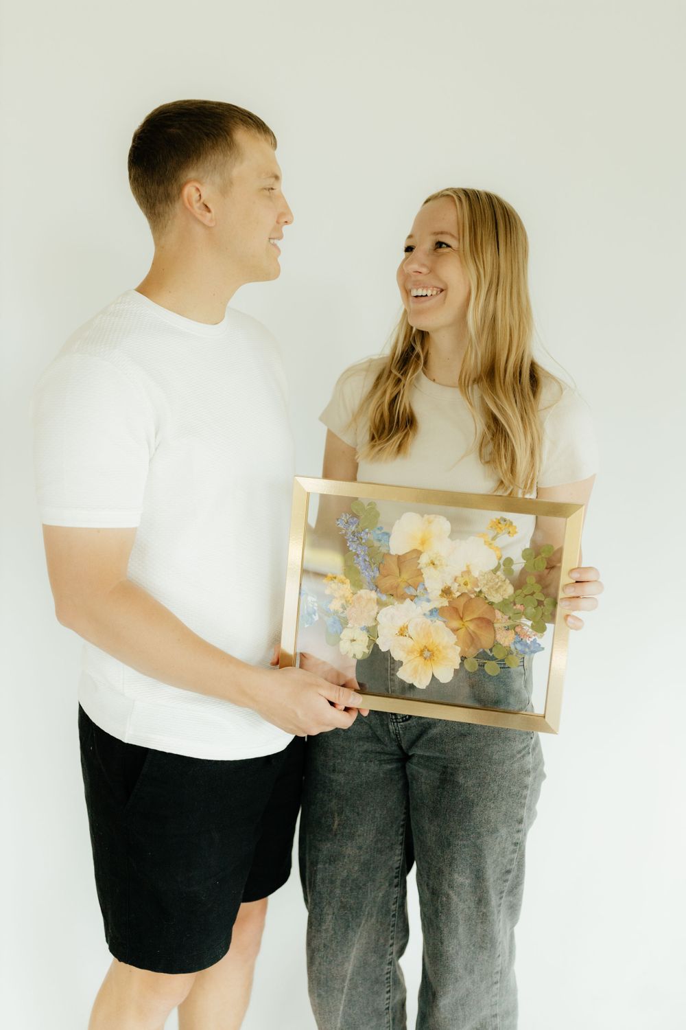 Smiling couple holding a gold-framed pressed flower artwork together against a white background.