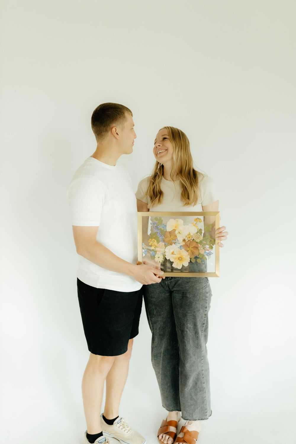 Smiling couple holding a gold frame with colorful pressed flowers against white background.